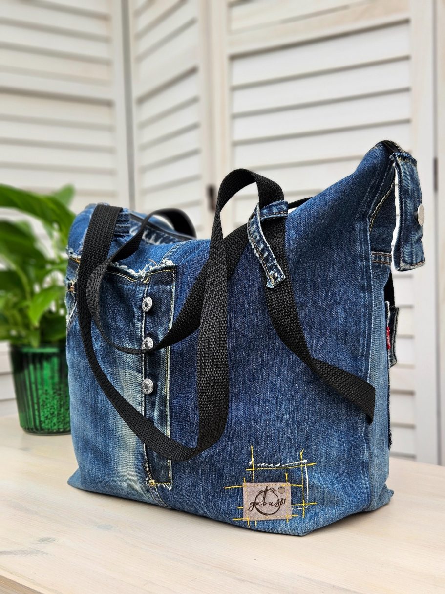 Denim shoulder bag with buttons and a black strap, next to a potted plant.