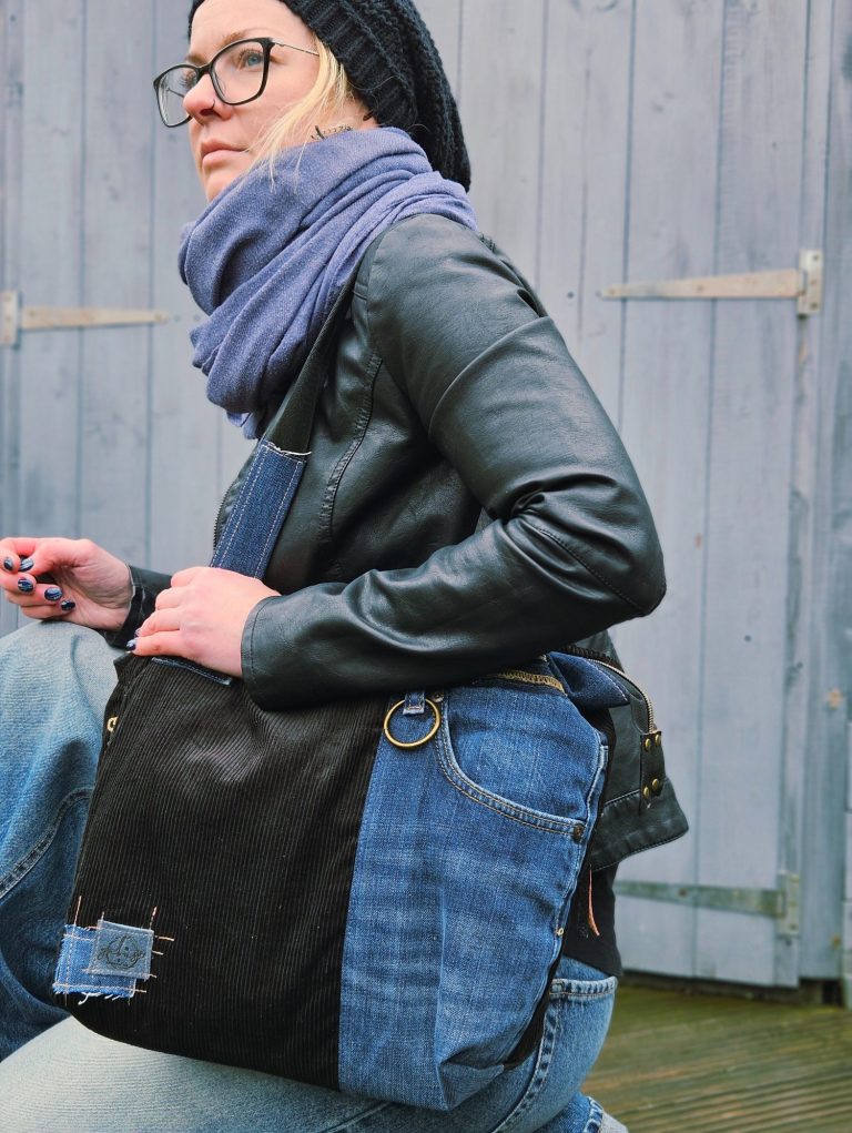 Person wearing a leather jacket and scarf, holding a denim bag, seated against a wooden backdrop.